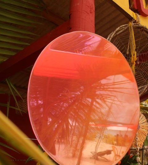Espelho redondo rosa pendurado num quiosque de praia, refletindo coqueiros.
