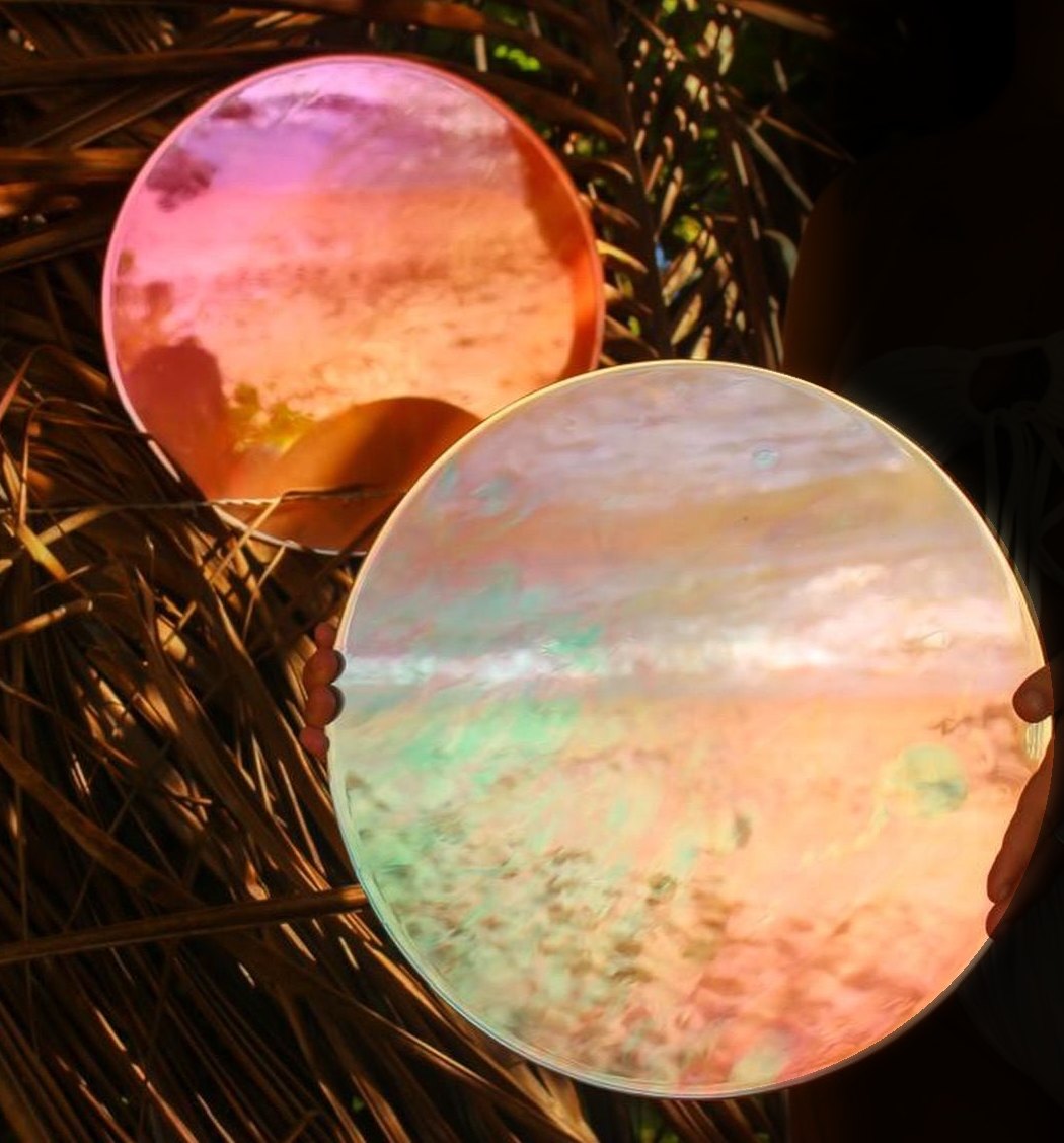 Dois espelhos redondos iridescentes, em rosa, laranja e verde, segurados entre folhas secas de palmeira.